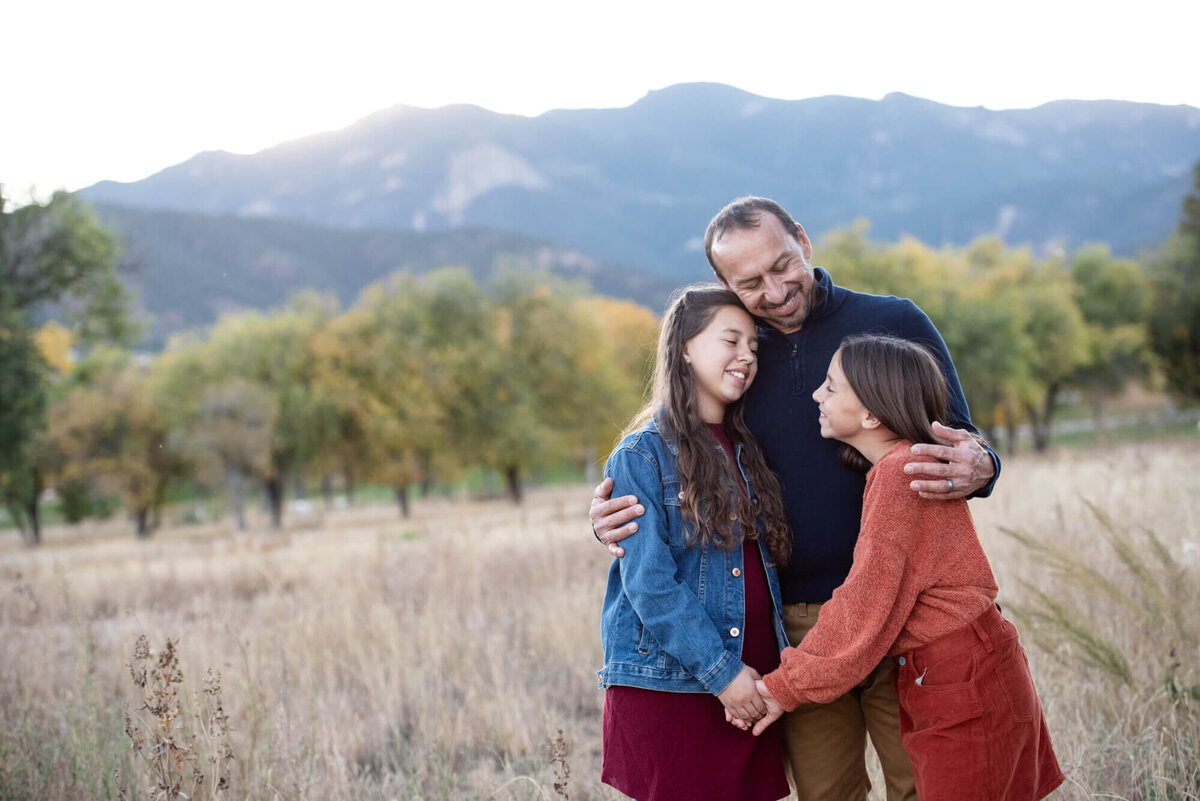 A father hugs his two young daughters while standing in a park field at sunset