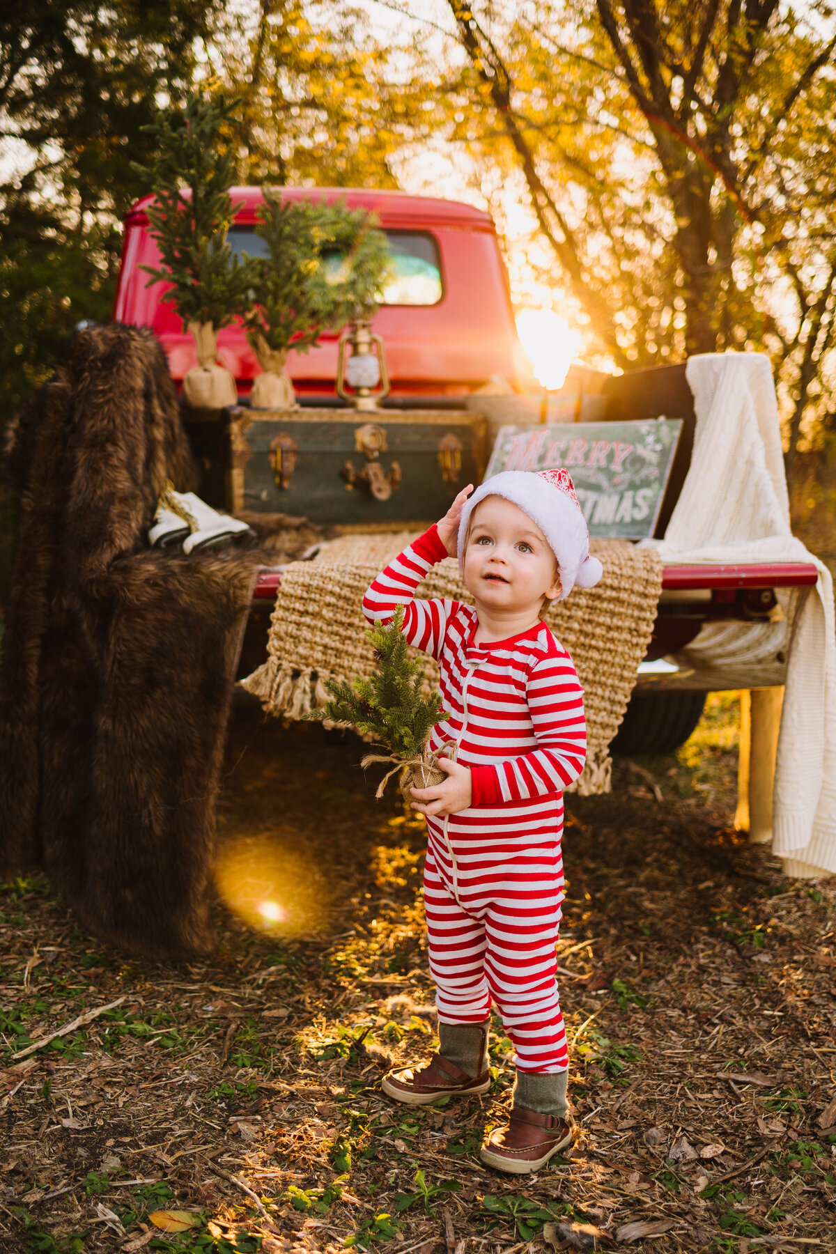 The baby boy is dressed with a red and white pajamas. He is in front of a red truck car and it is decorated with christmas decoration.