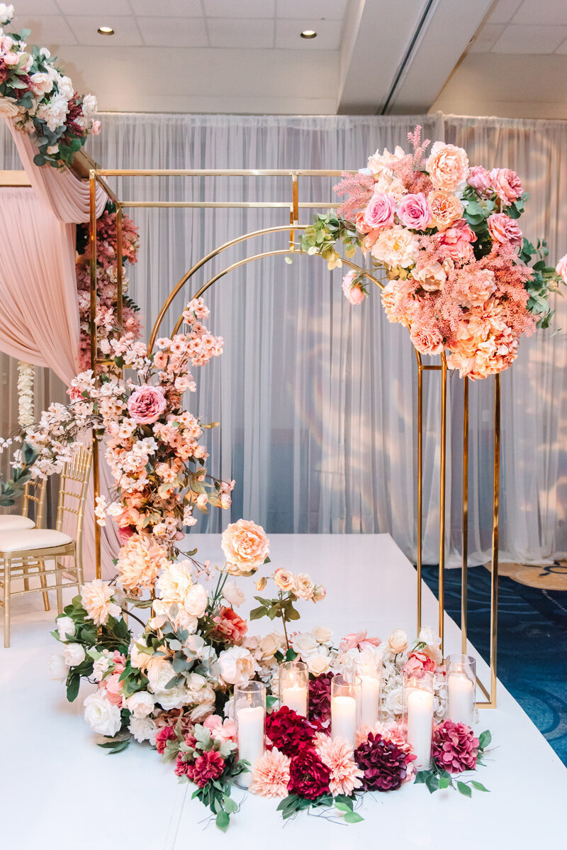 Wedding decor that features a gold arch  with pink florals and tall white candles.