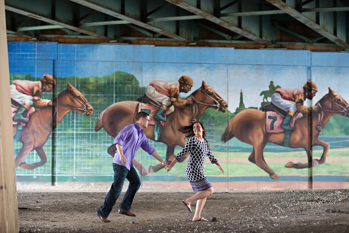 A bride to be runs with her fiance in front of a mural of a horse race in northern liberties.