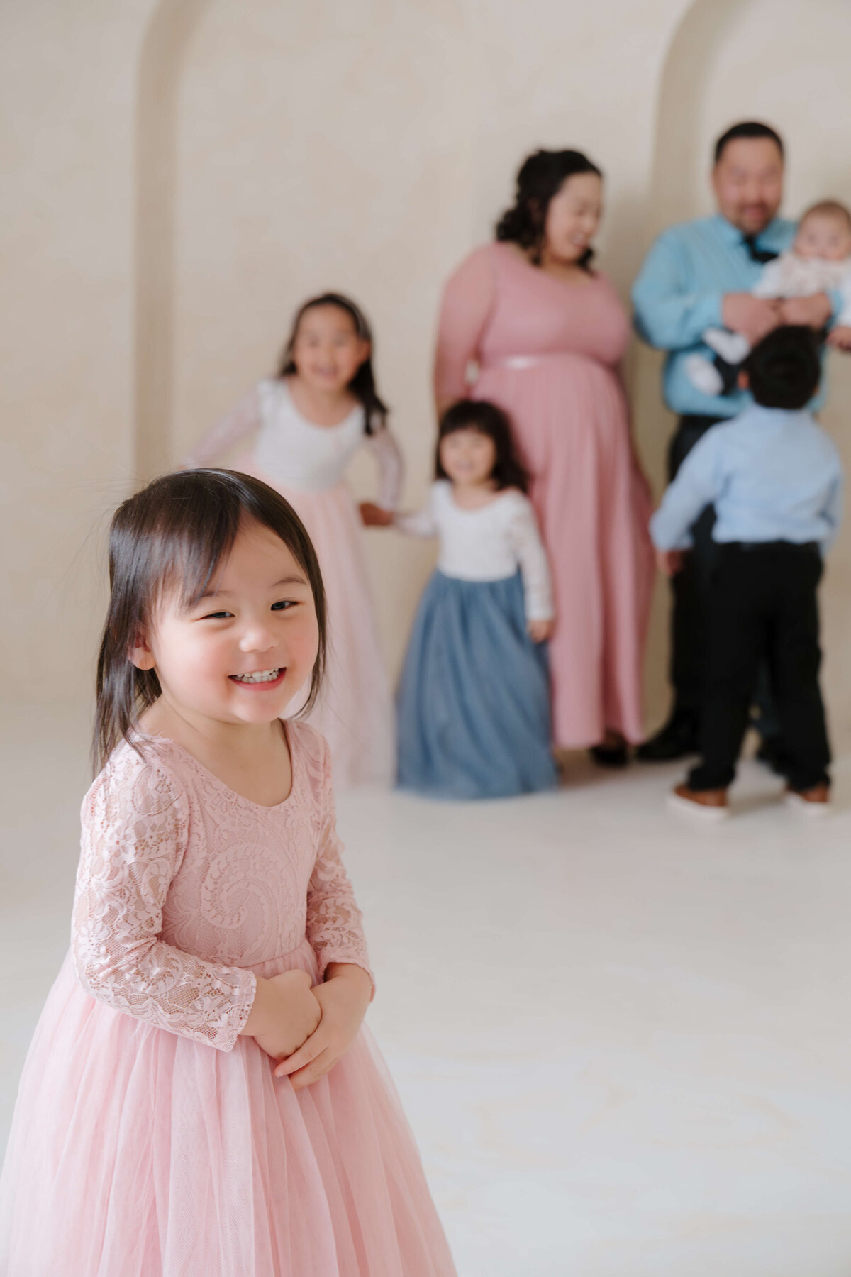 Young girl in a pink lace dress smiling in the foreground with her family blurred behind her.
