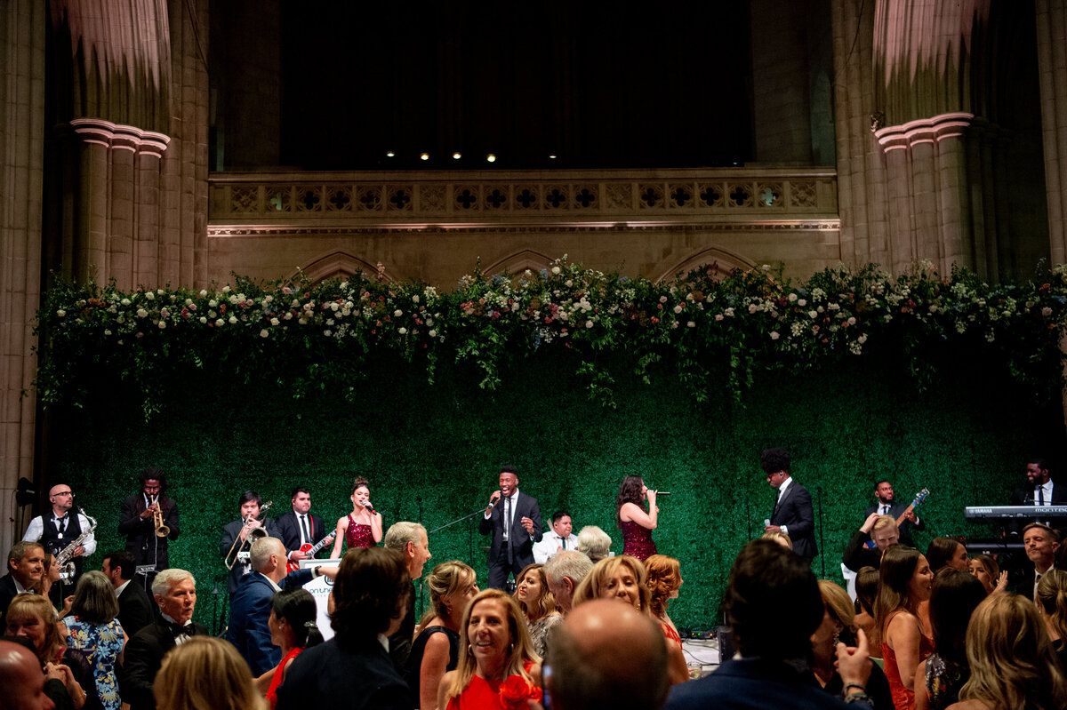 This lively scene unfolds at the Washington National Cathedral in Washington D.C., captured by the talented photographer Twah Dougherty. A band performs energetically on stage within a large hall featuring high ceilings and intricate architectural elements. The backdrop is enhanced with lush greenery and beautiful flowers, adding to the ambiance. In the foreground, a vibrant crowd of individuals, elegantly dressed, sways to the music and revels in the enchanting atmosphere.