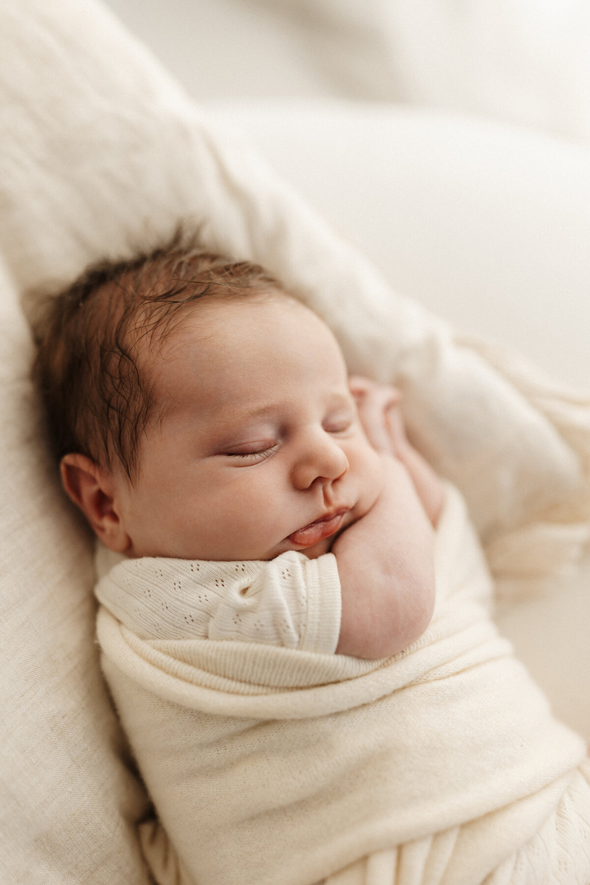 A newborn baby with dark brown hair and cute chubby cheeks wrapped in a white blanket and sleeping during his newborn photoshoot with popular newborn photographer lucy jane