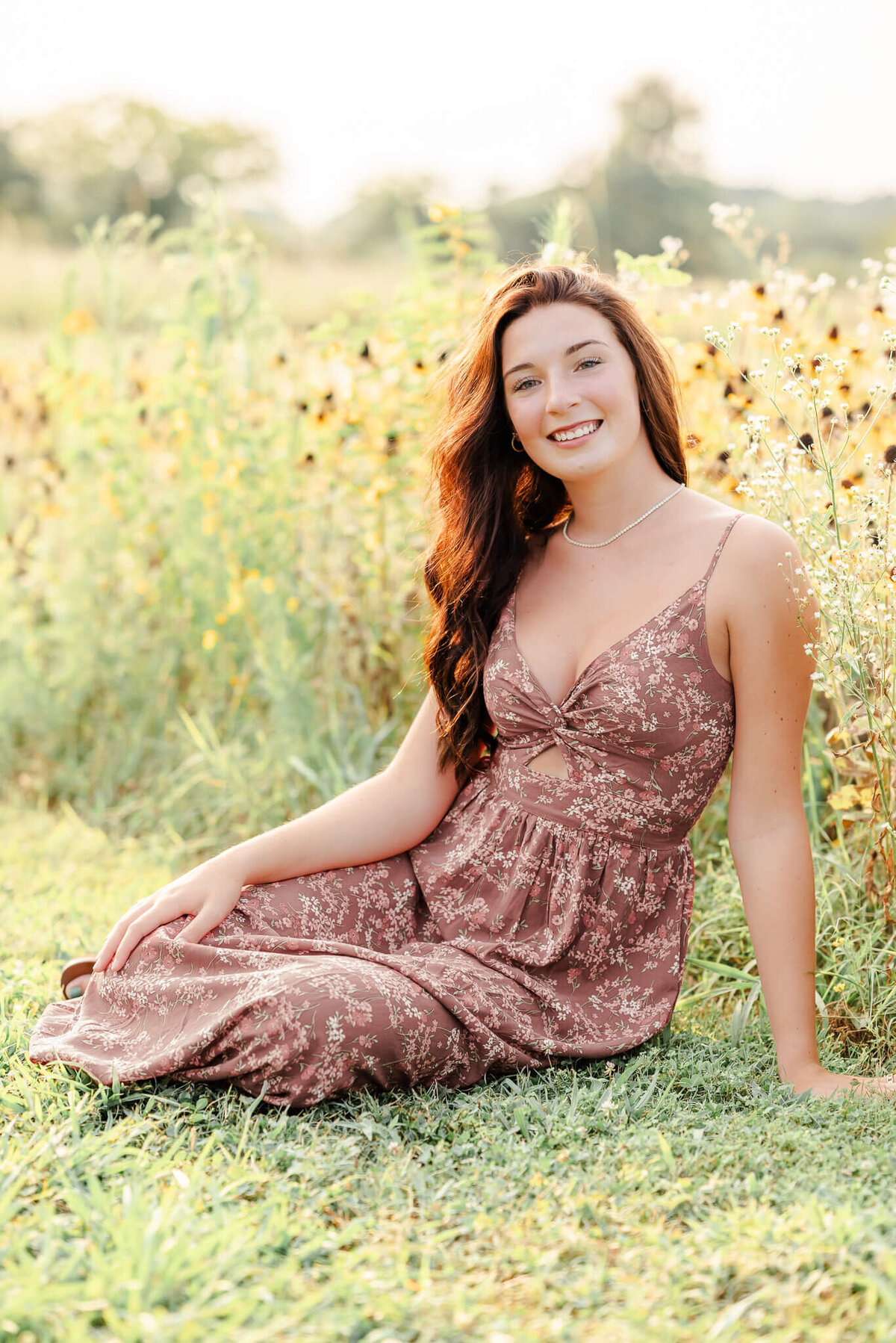 A high school senior in a floral dress sits in front of some yellow flowers. She is having her senior session with Chesapeake Photographer, Justine Renee Photography.