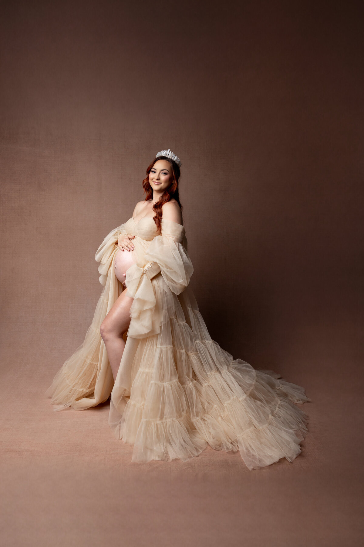 Wearing a dusty white dress and a delicate crown, she smiles softly while cradling her belly, her serene expression highlighted by gentle lighting. Captured by Inland Empire photographer Kaitlyn Dawn Photography.