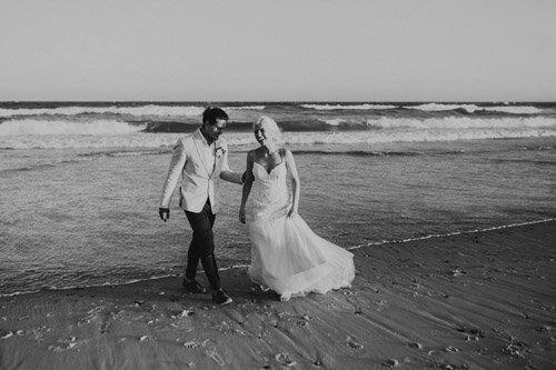 B+W photograph of a beach wedding on the NSW Central Coast at Caves Beach. The couple are walking in the surf and laughing.