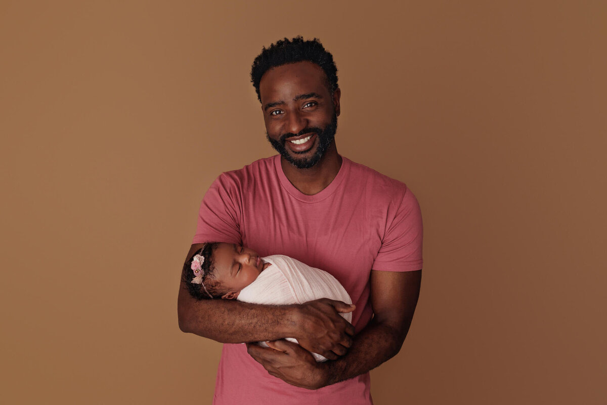 A smiling father in a pink shirt holds his newborn baby, swaddled in white, against a brown background.