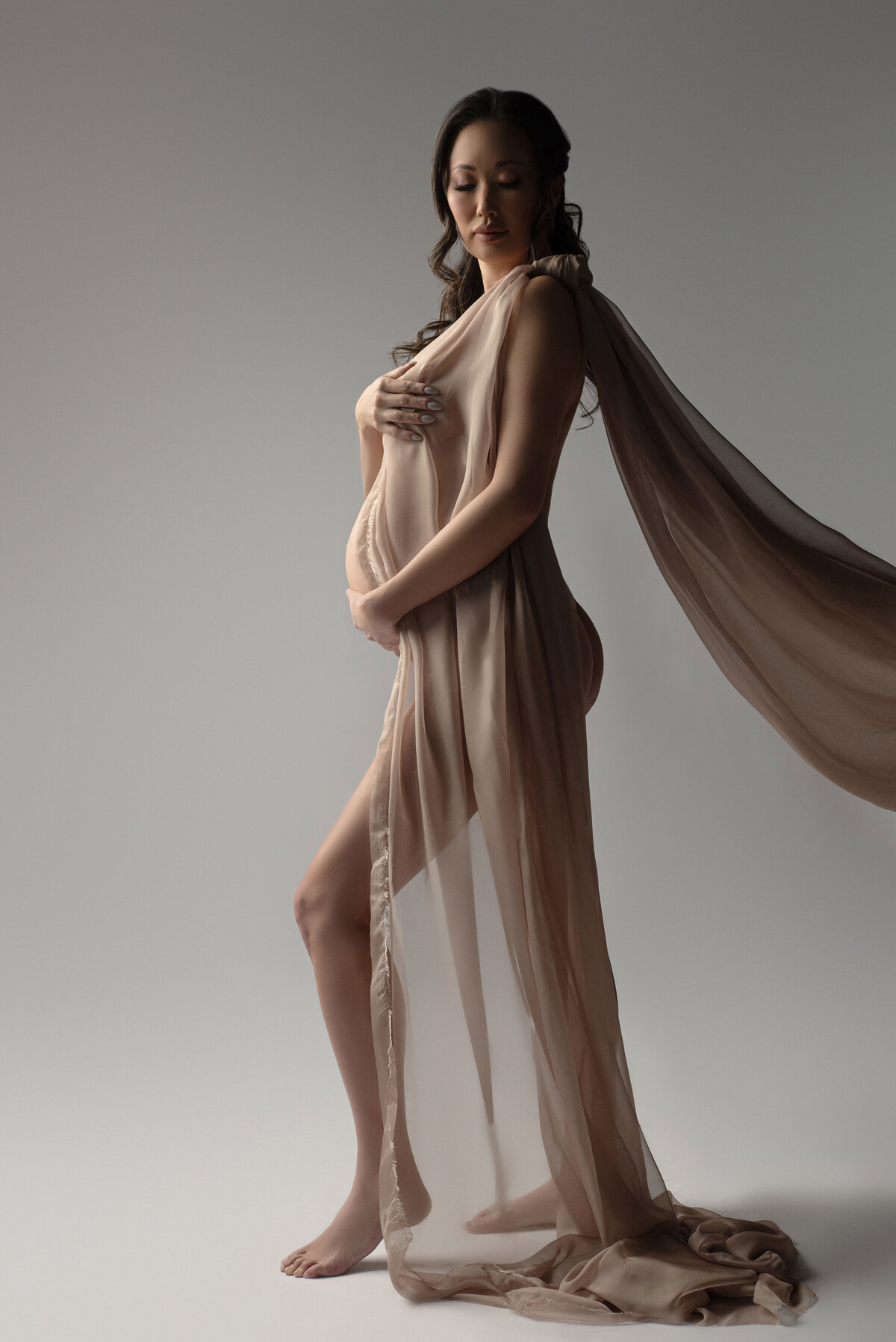 pregnant woman draped in beige fabric with  backlit lighting and holding belly looking down pose