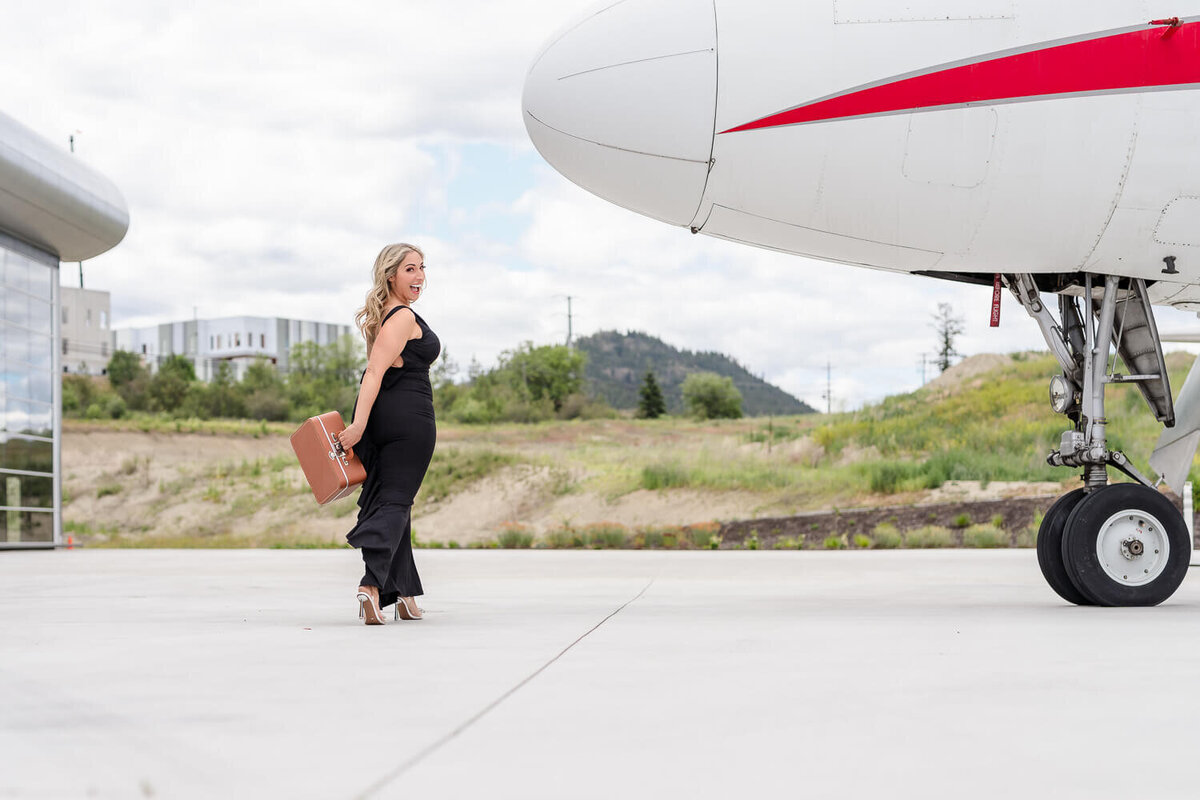 Female jet setter in black dress walking with vintage suitcase toward an airplane in Kelowna.