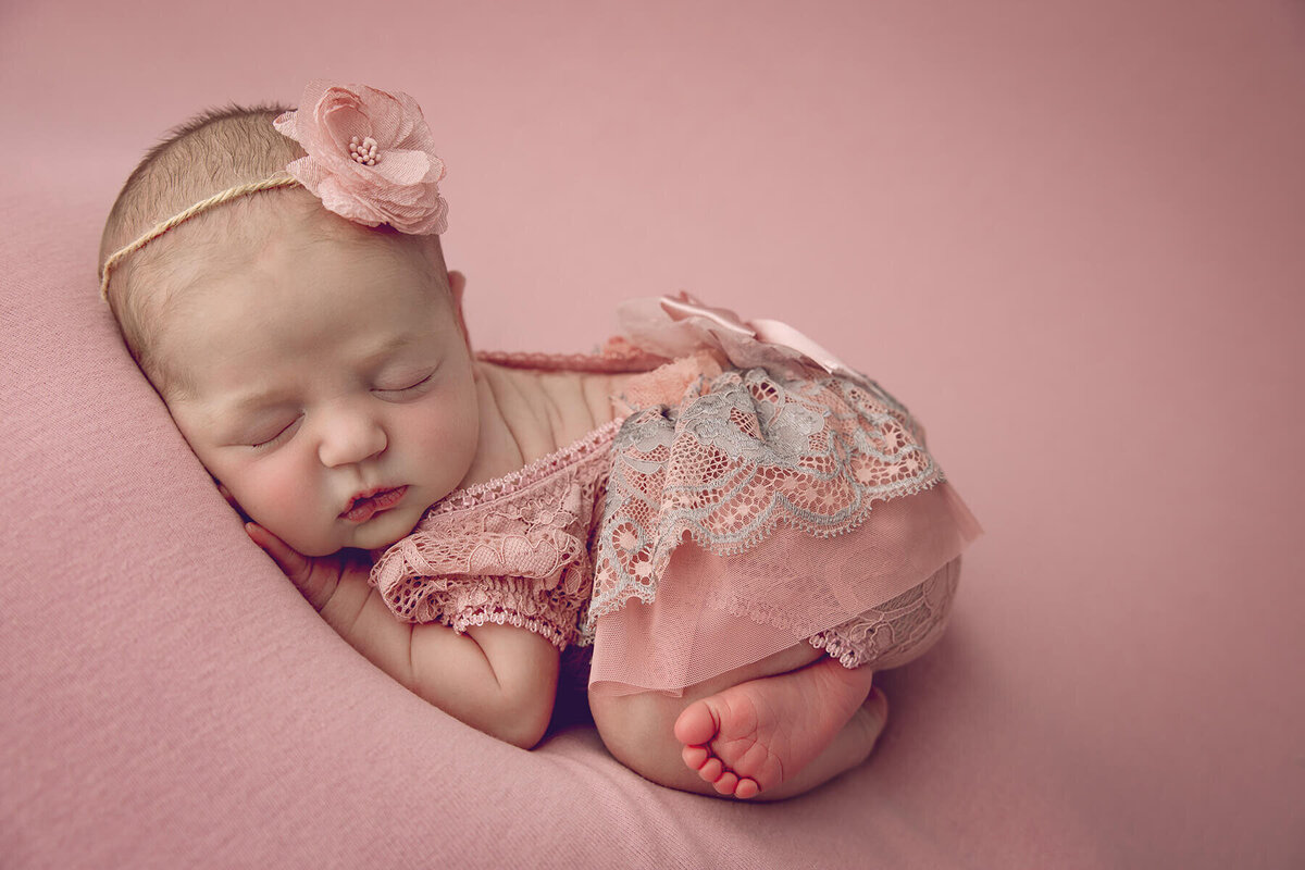 Baby girl in pink lace outfit sleeps curled up on a matching blush backdrop with floral headband.