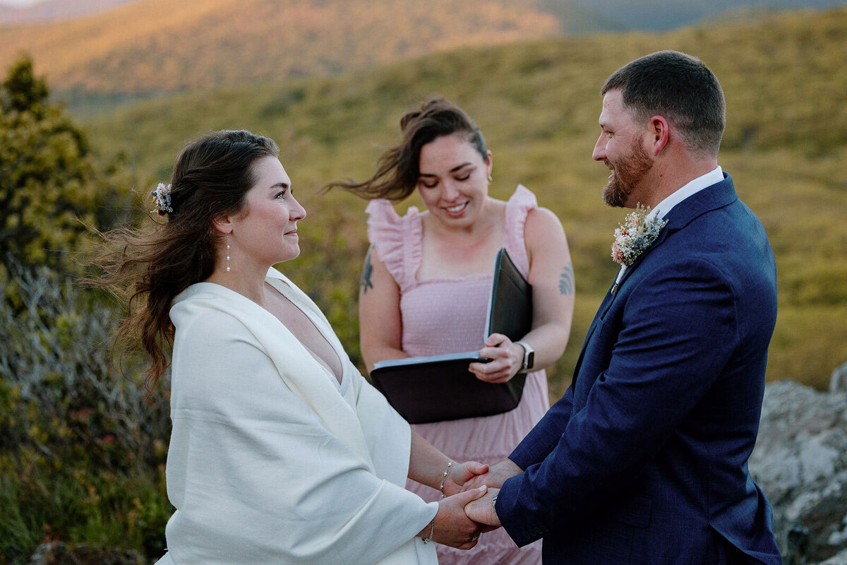 A bride and groom look at one another during their vows on their wedding day. They are holding hands, and the sun is rising over the mountains behind them. 