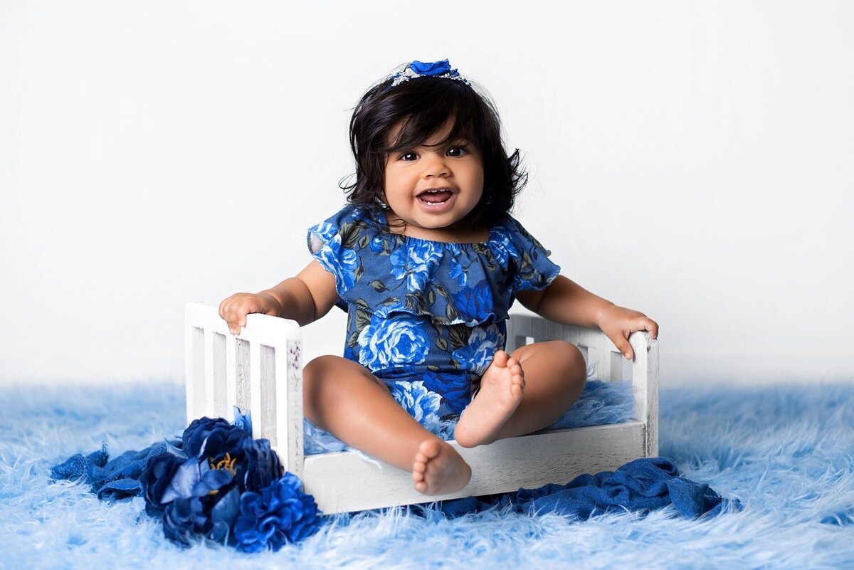 Baby girl in blue floral romper laughing on white prop bed.