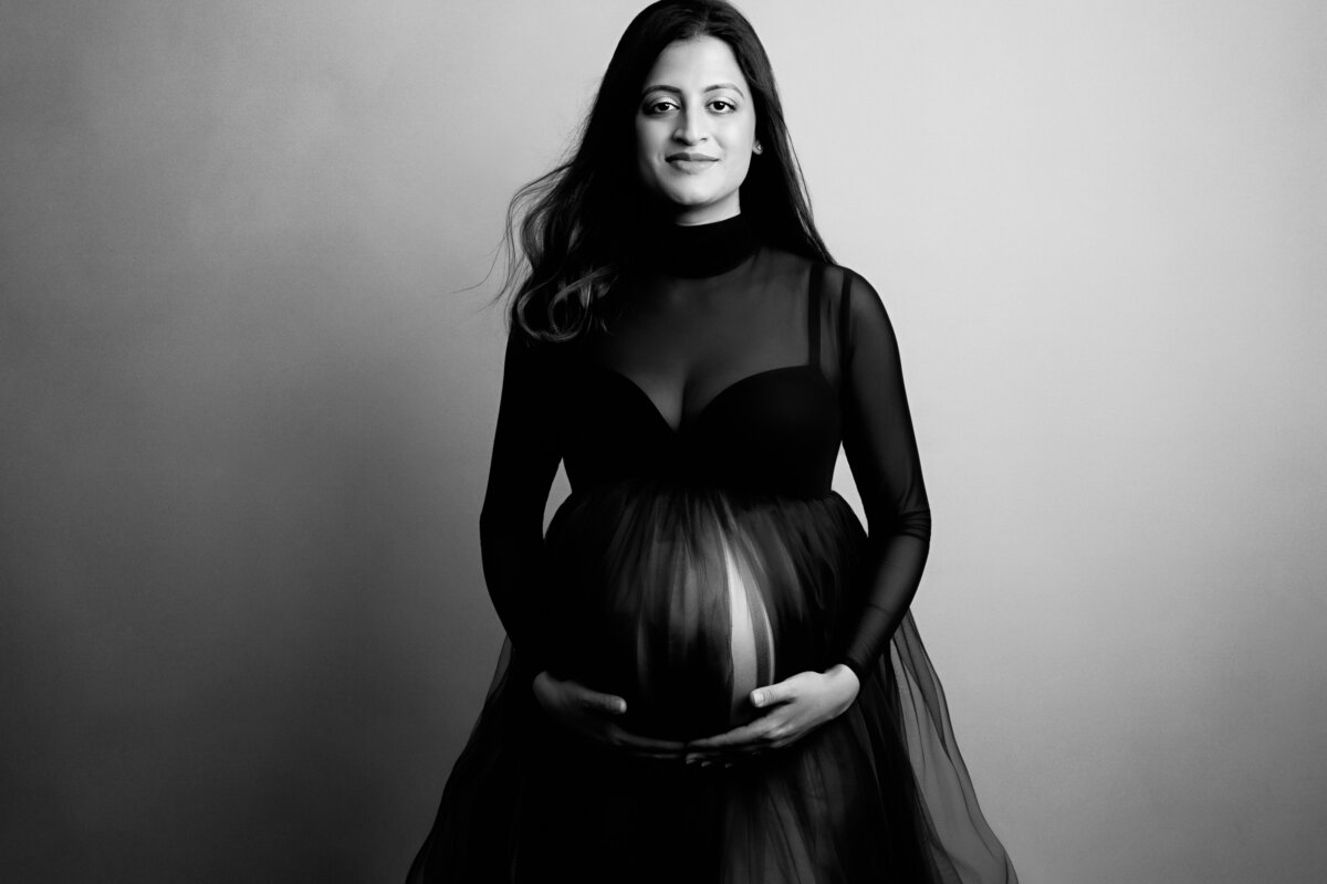 Classic style maternity portrait of a woman wearing a sheer long black dress with her hands under her belly.