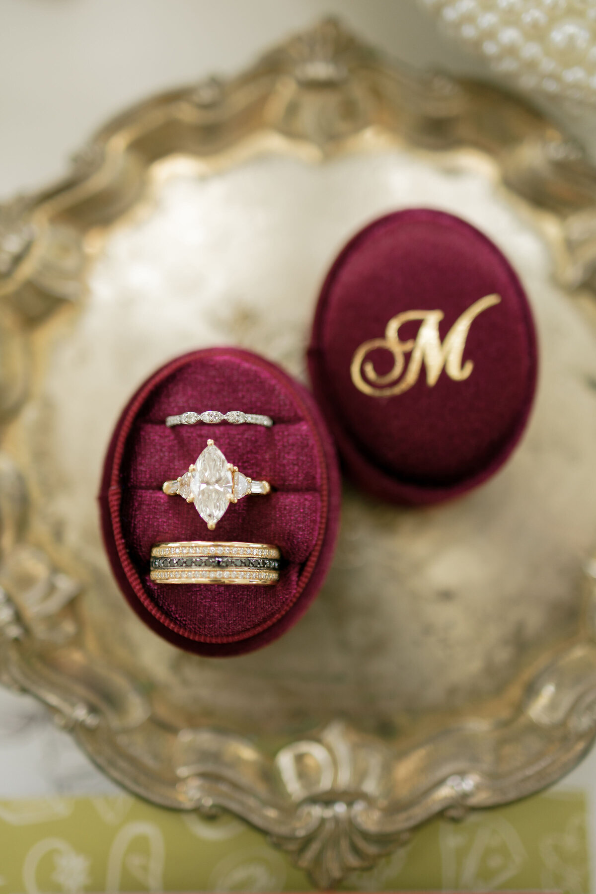 Deep burgundy velvet ring box styled on an ornate vintage silver tray, holding a marquise-cut diamond engagement ring with side stones and two coordinating wedding bands, captured as elegant flat-lay wedding details.