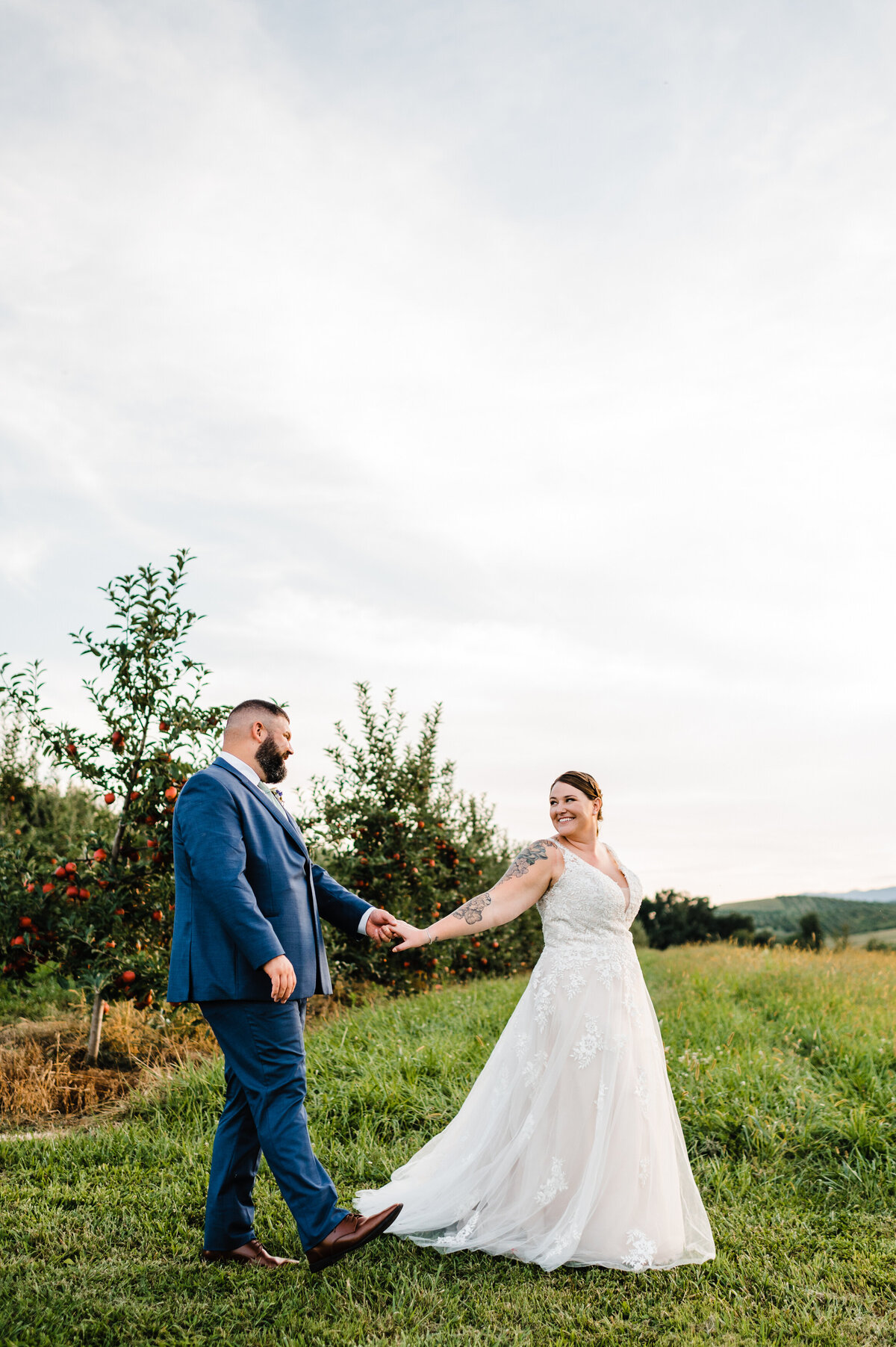 showalter's orchard wedding with bride and groom walking through an apple orchard together while holding hands and the bride looking back at the groom as they exit the orchard and walk into a field