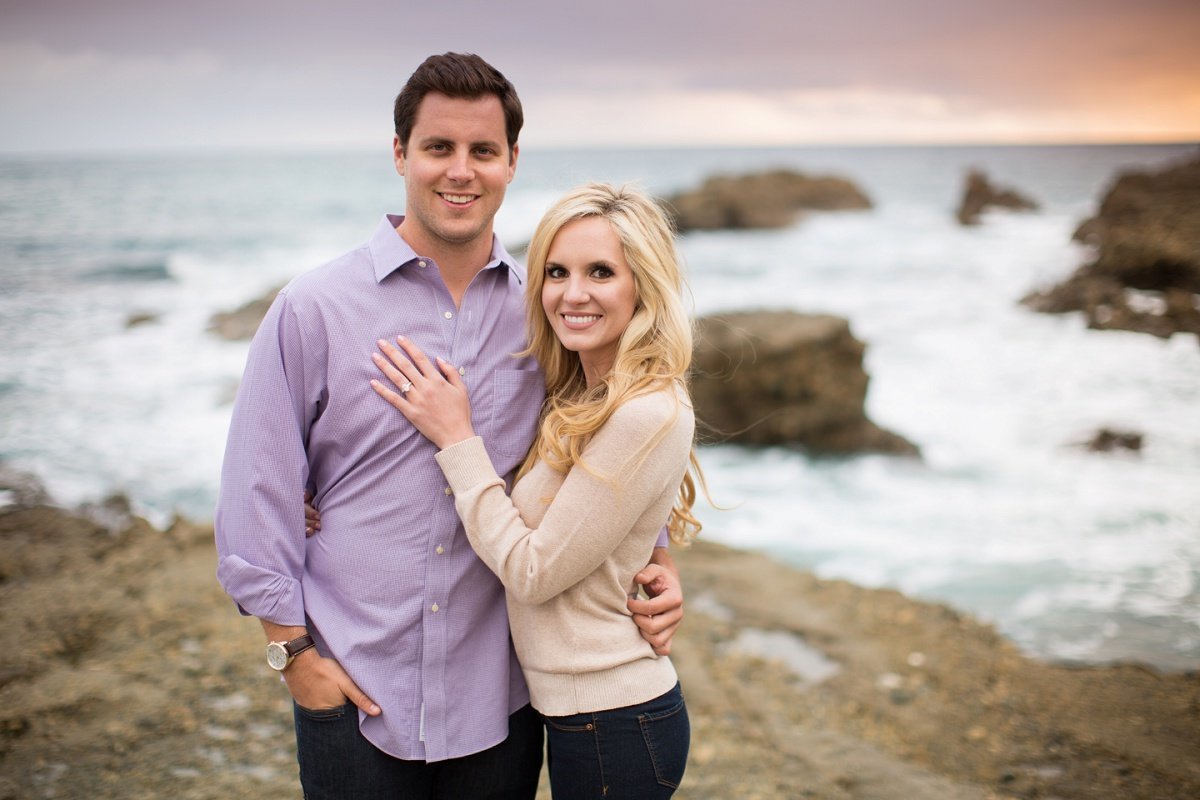 Engaged couple cozy up to each other as they pose for engagement photos on a cliff overlooking the ocean waters crashing up against large rocks