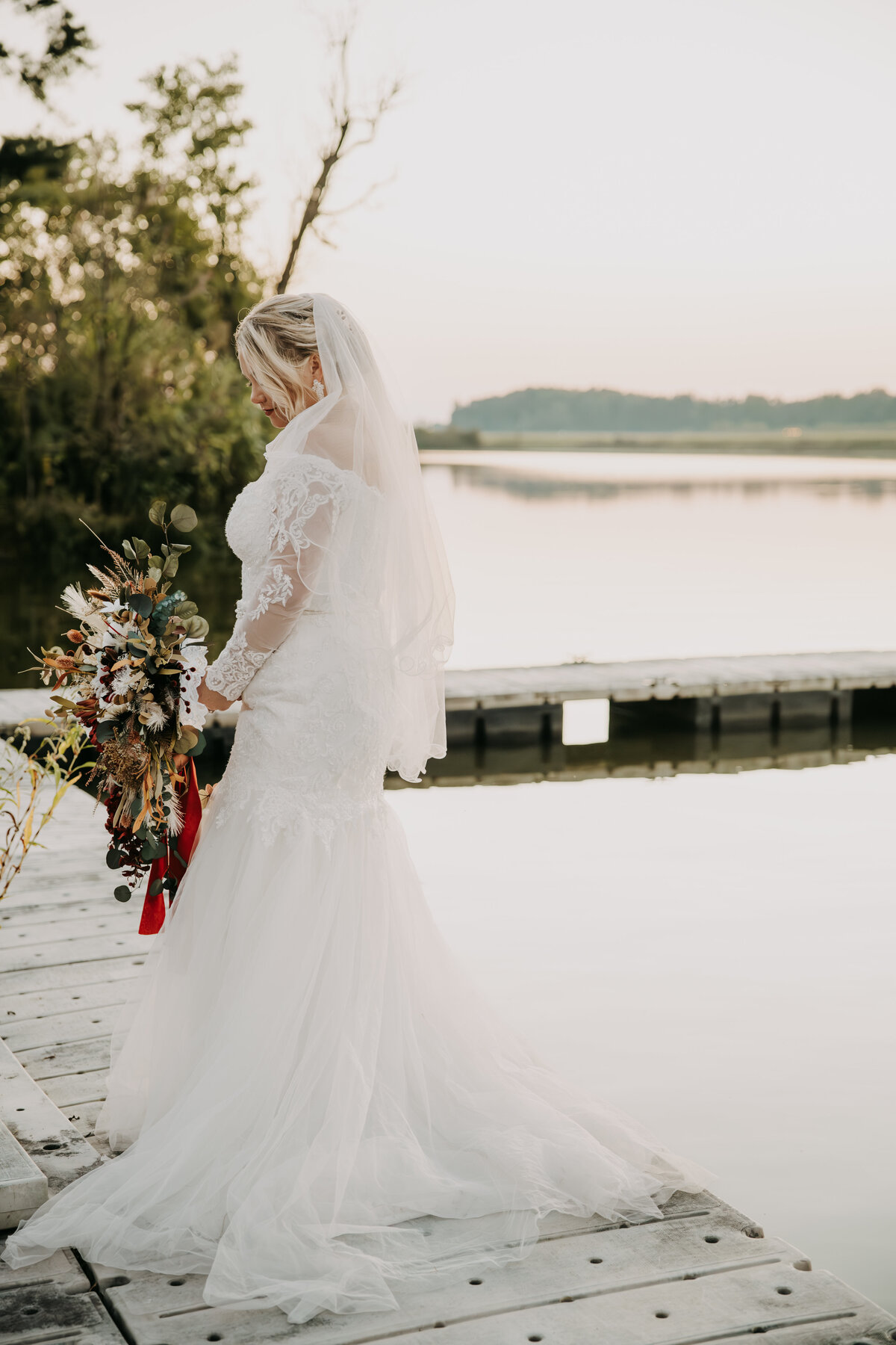Bride standing on a dock with her back to the camera, holding a bouquet of fall-colored flowers, her dress train trailing behind her, illuminated by the soft glow of sunset.