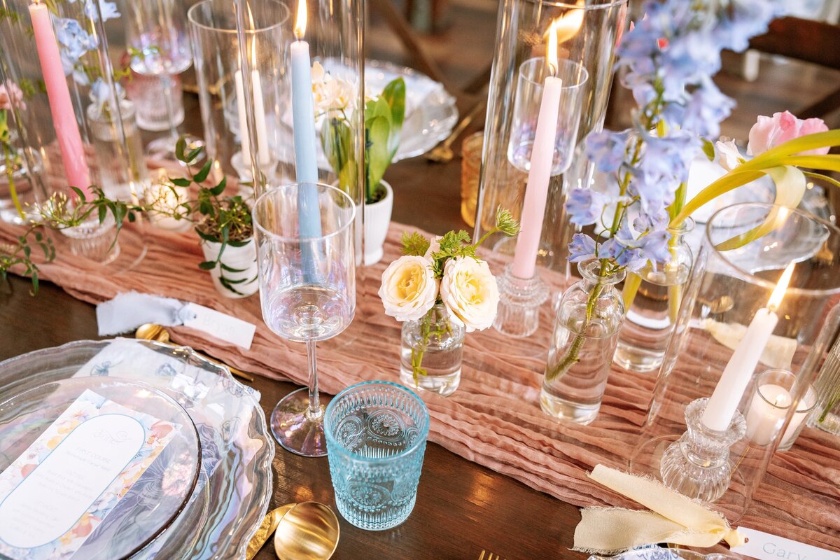 A beautifully set table at an exclusive Leesburg, VA wedding venue features floral arrangements, tall candles in glass holders, and exquisite glassware. The centerpiece boasts light blue and purple flowers, complemented by elegant gold cutlery and delicate white rose accents.