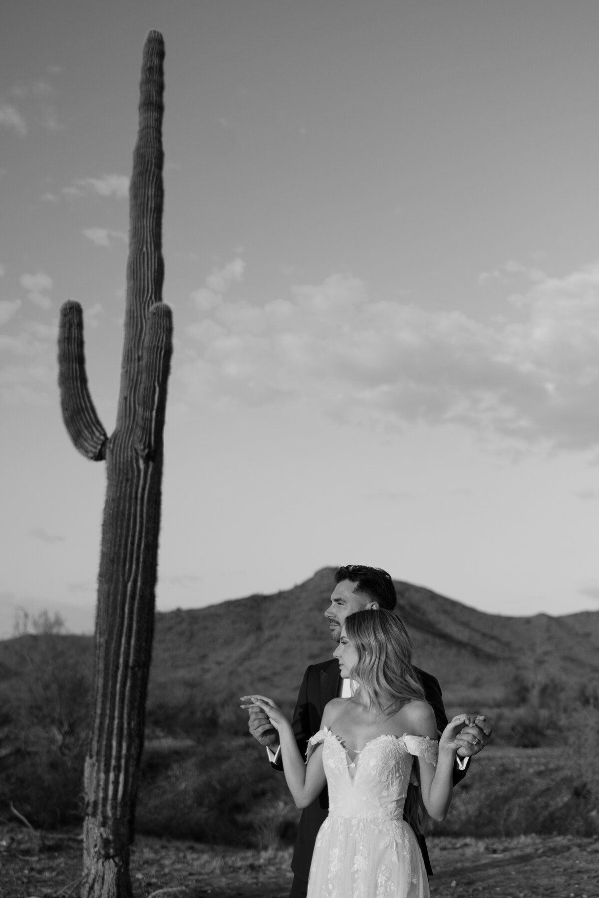 a bride stands in front of her groom as he holds her hands to the side with a cactus to the side of them