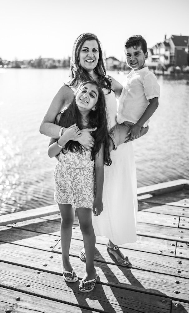A mom in a white dress is holding her son on her hip and hugging her daughter with her other hand