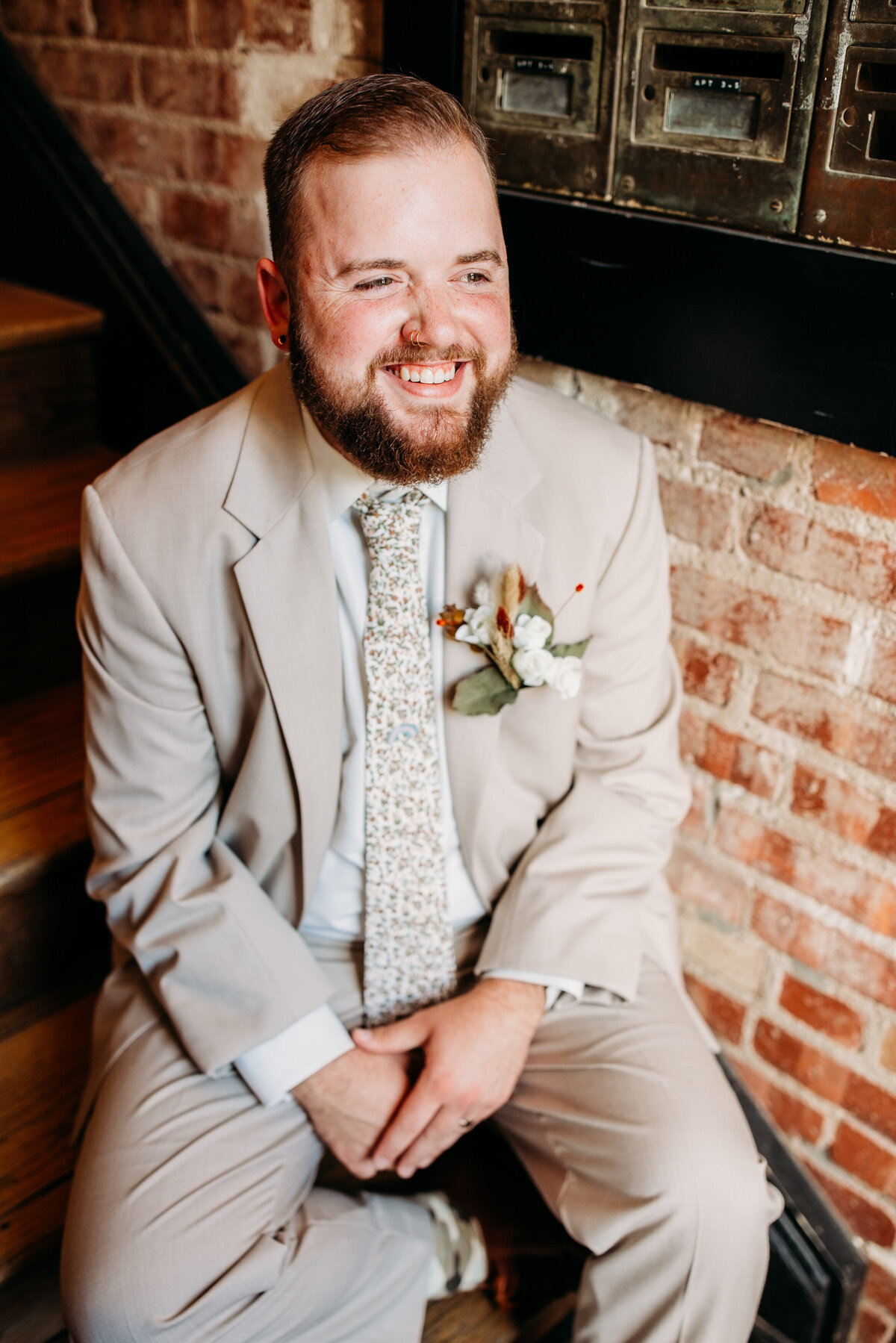 Groom seated and smiling broadly against a brick wall, wearing a beige suit and a floral tie.