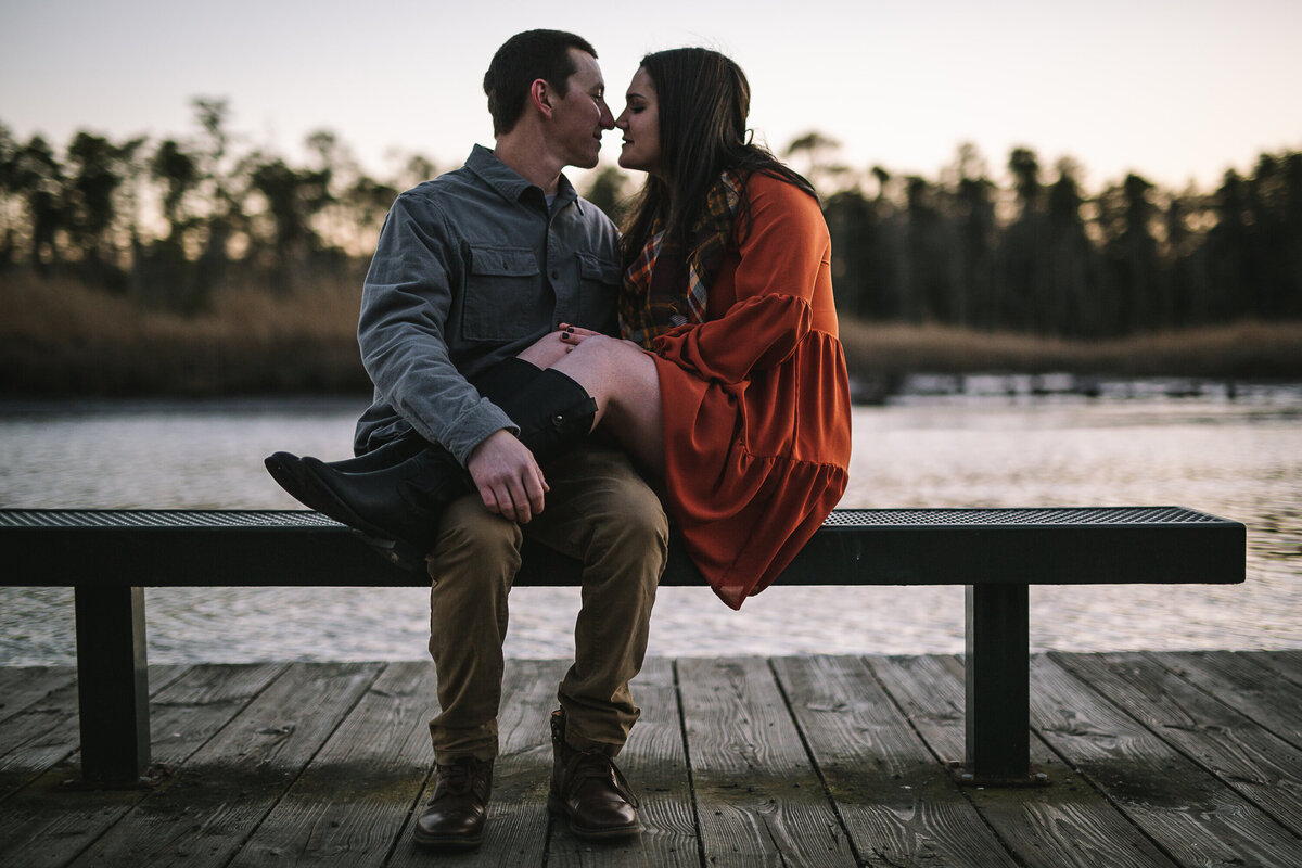 Woman has her legs over her fiance's lap on a bench by the water.