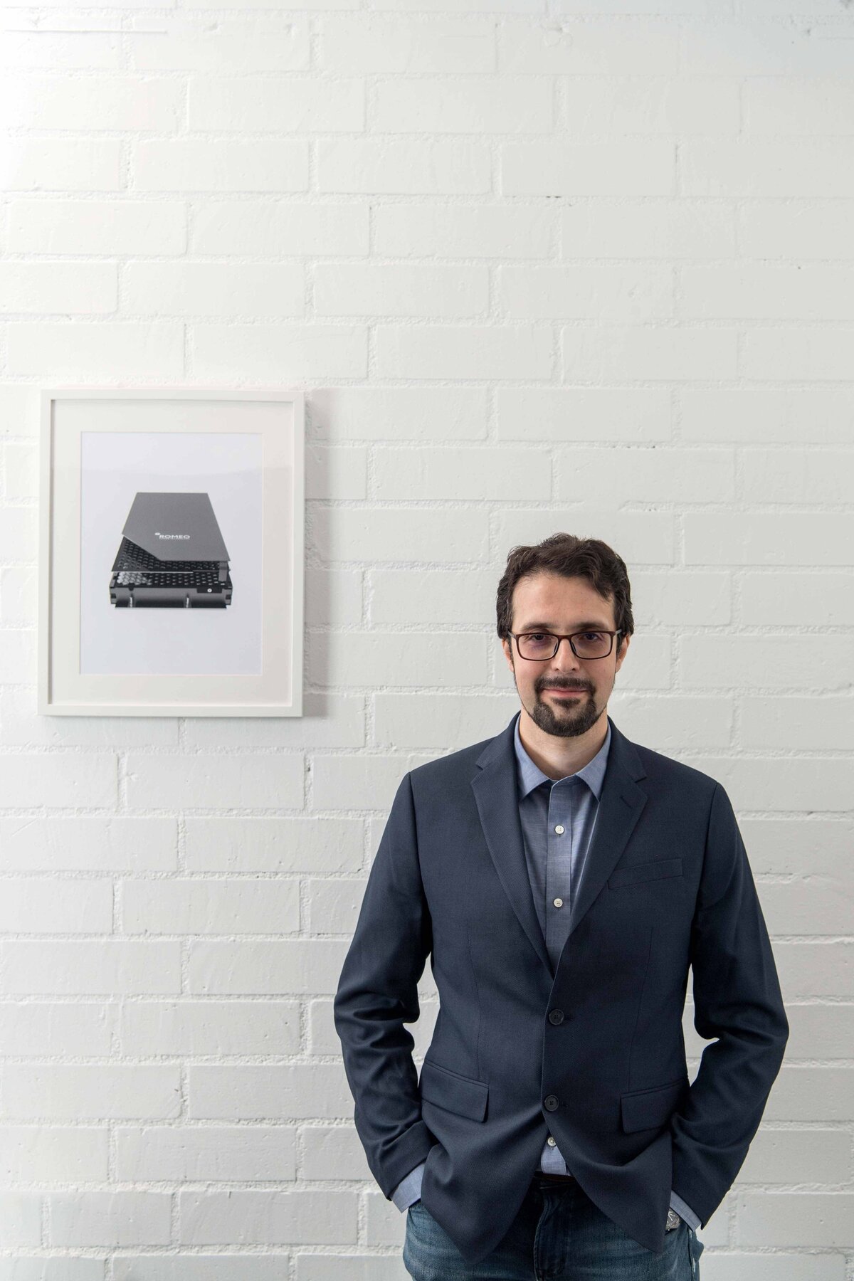 Editorial Headshot of a man with suit and glasses standing against white brick wall with image of his invention behind him