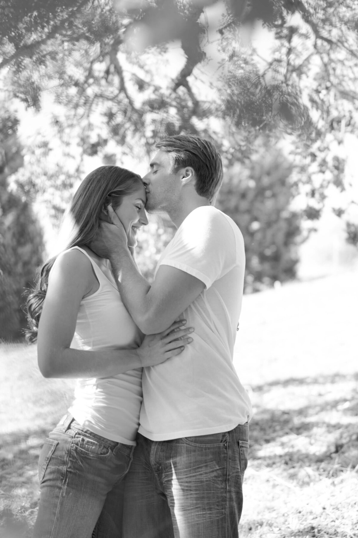 Newly engaged couple in jeans and white tshirts celebrate their casual engagement outdoors with a kiss on the forehead.