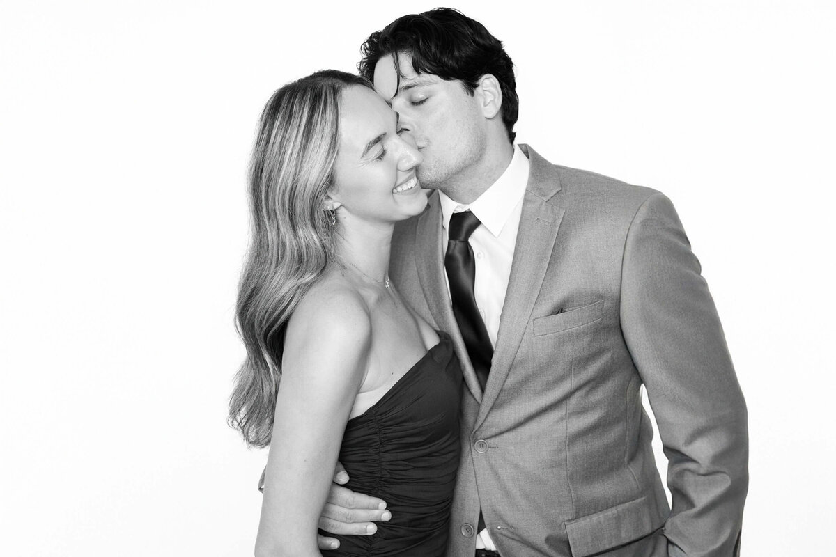Couple sharing a joyful moment as he kisses her cheek in a black and white portrait by Monochrome Portraits, a luxury Phoenix photo booth experience.