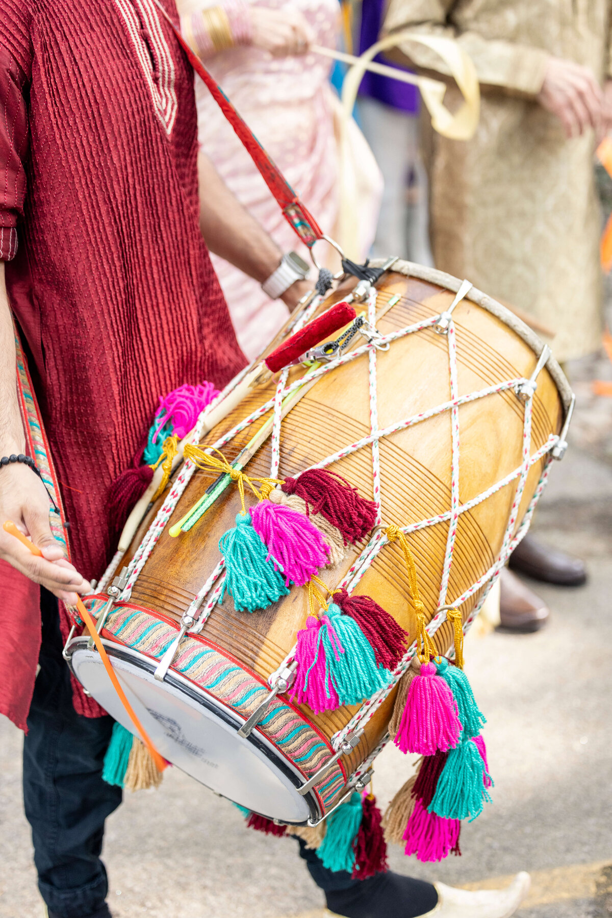 A person wearing a maroon kurta plays a traditional dhol drum adorned with pink and turquoise tassels. The background shows other people in colorful attire, suggesting a festive or cultural event.