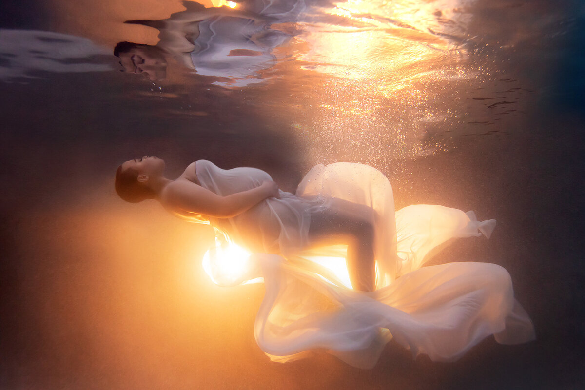 A woman gracefully floating underwater in a sheer white gown, illuminated by a warm, glowing light from below. The ethereal scene captures tranquility and elegance, with golden light reflecting off the water's surface and creating a dreamy, surreal ambiance.
