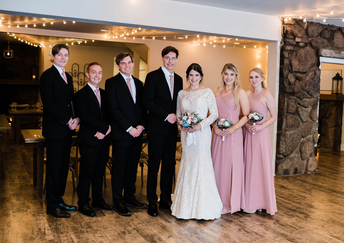 Wedding party consisting of 2 bridesmaids in pink short sleeve dress and groomsmen in black suits stand in a line with the bride and groom in the middle smiling for the camera