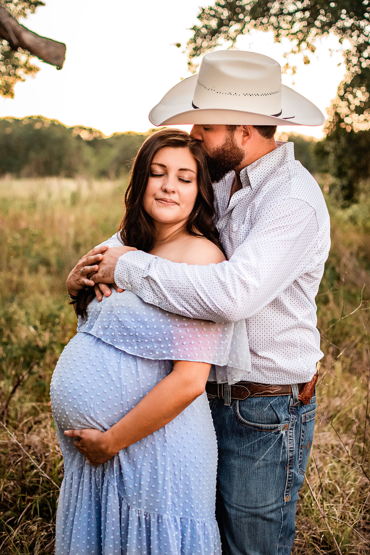 A husband hugs his pregnant wife from behind while he kisses her forehead.