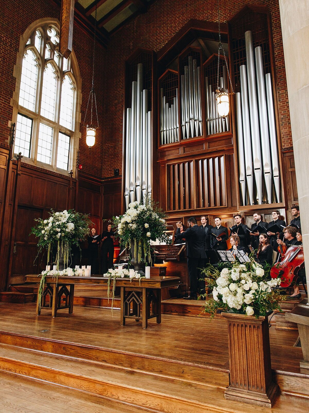 Orchestra plays music for a wedding at Scarritt Bennett on the church alter next to the large pip organ. The altar is decorated with  lots of white candles and tall floral arrangements with hydrangea, white roses and greenery.