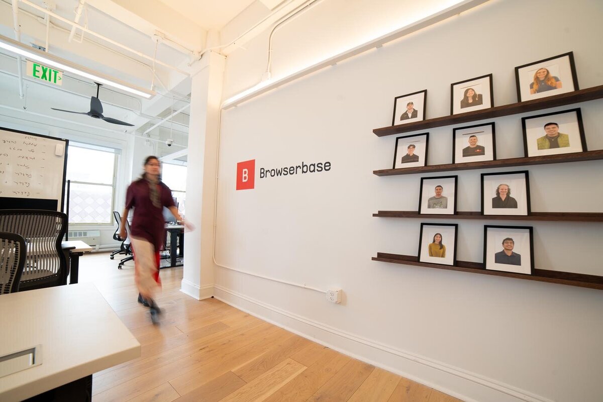 Office hallway with a “Browserbase” logo on the wall, framed employee portraits on floating shelves, and a person walking past in motion blur.