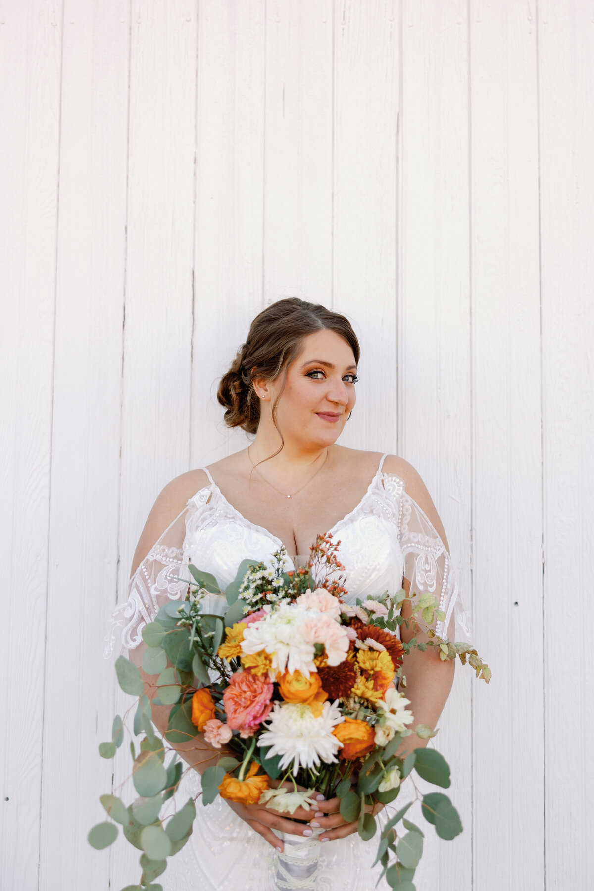 Bride in lace gown holding a vibrant autumn bouquet of orange, white, and rust flowers outside The Hummingbird Chateau in Manchester, Maryland.
