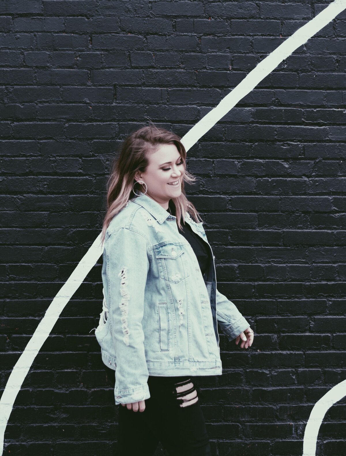 Woman in distressed jean jacket with black brick background.