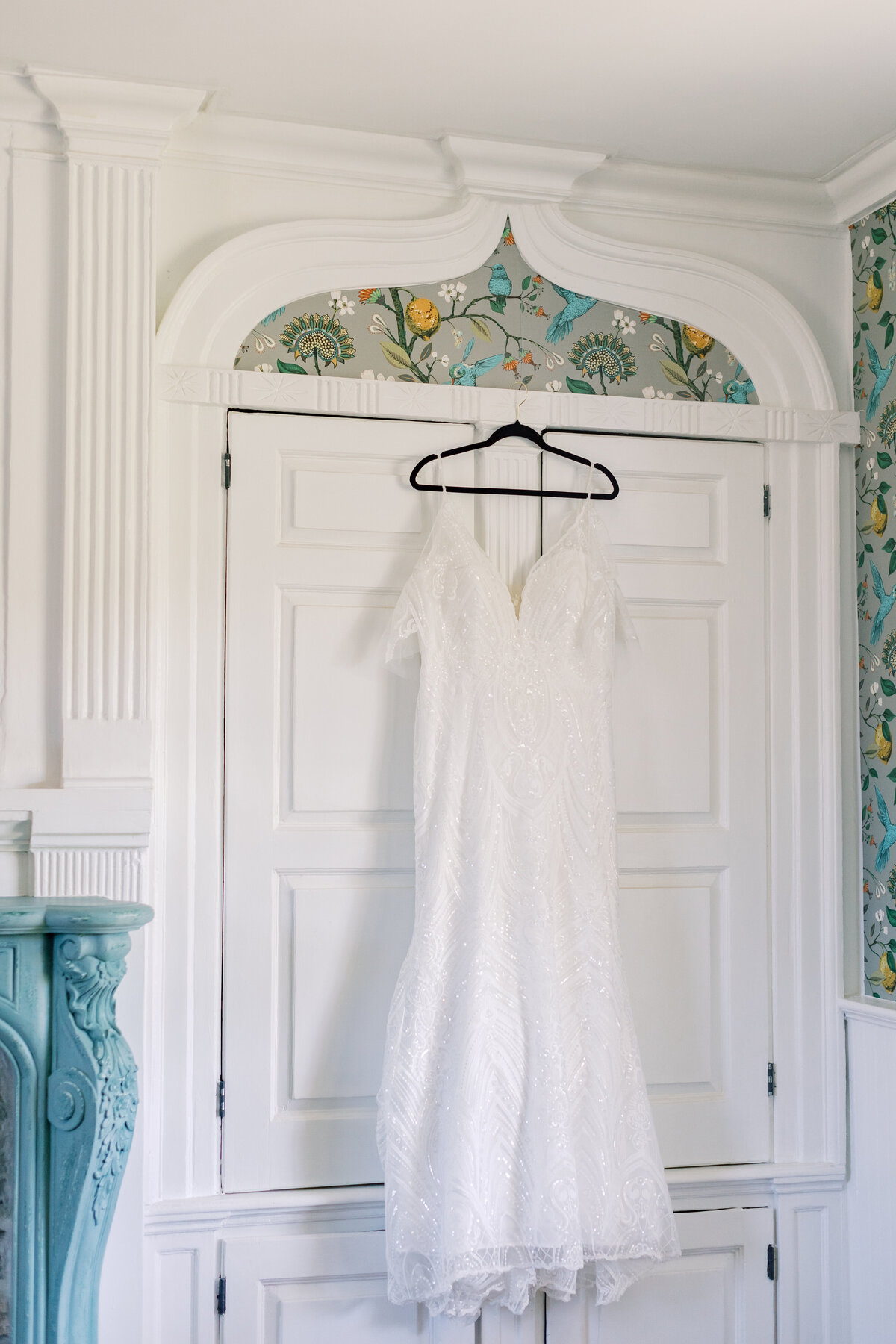 Wedding dress hanging on ornate white wardrobe with floral wallpaper accent.