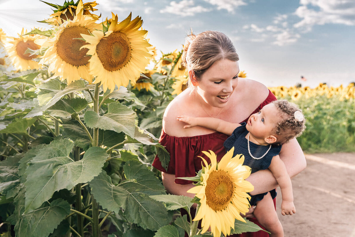 A baby girl reaches for the bloom in a sunflower field, while her mother holds her and smiles down at her.