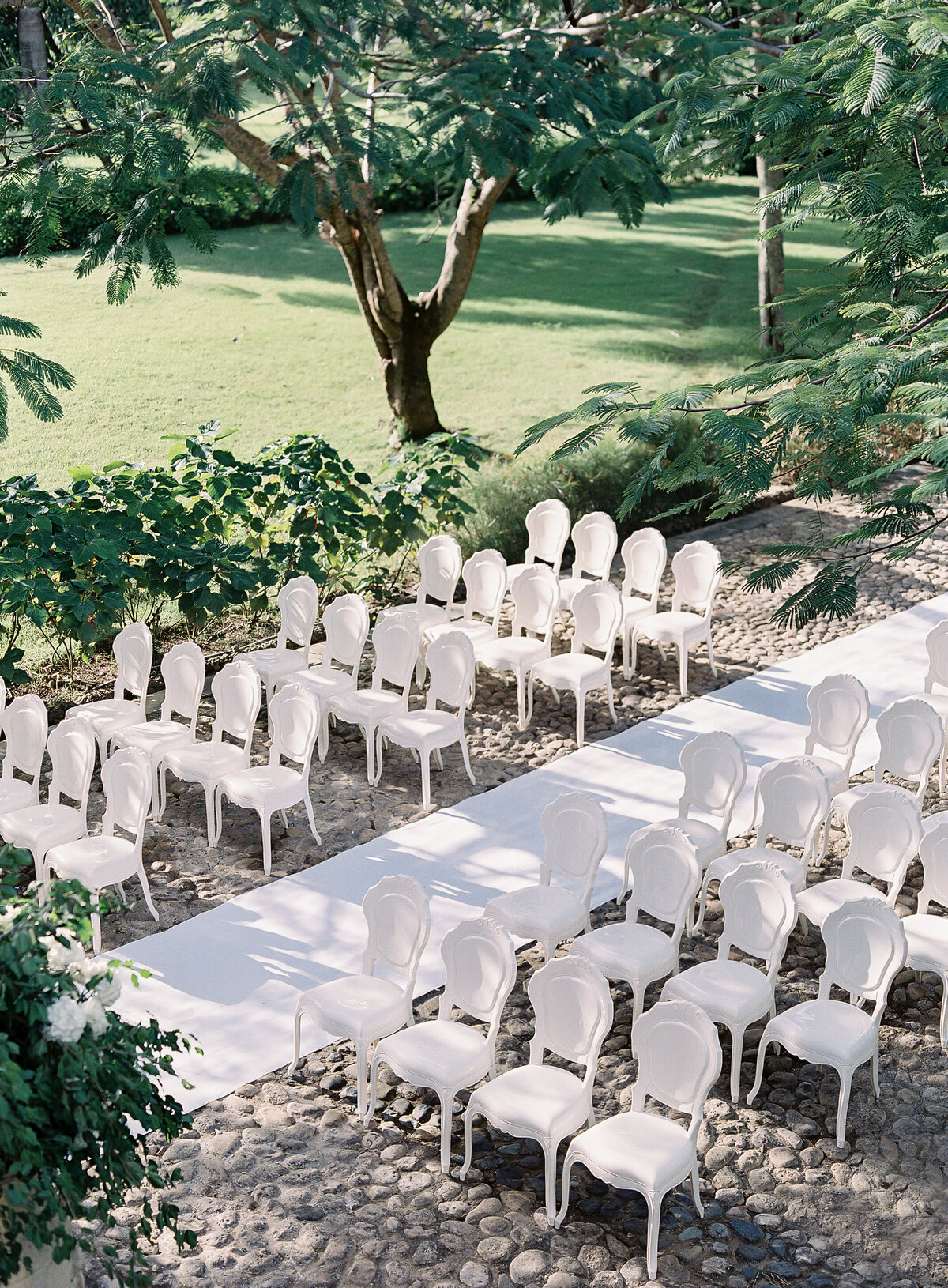 Outdoor wedding setup at a top tropical destination wedding venue in the Caribbean, Dominican Republic. Rows of white chairs arranged on pebble stone ground, surrounded by lush green trees and foliage. A white aisle runner leads to the front for the ceremony, captured beautifully by Photographer Laura Gordon.