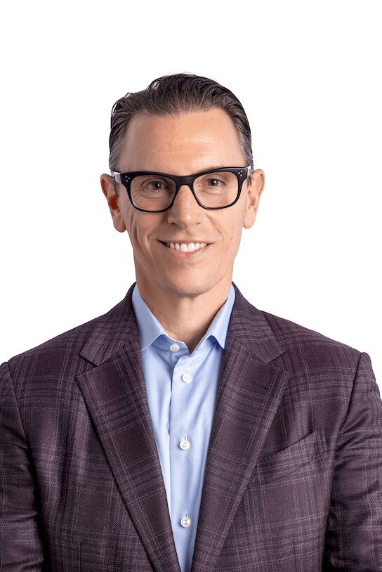 Real Estate Headshot of a man wearing a suit and eye glasses without a tie on a white background smiling at the camera