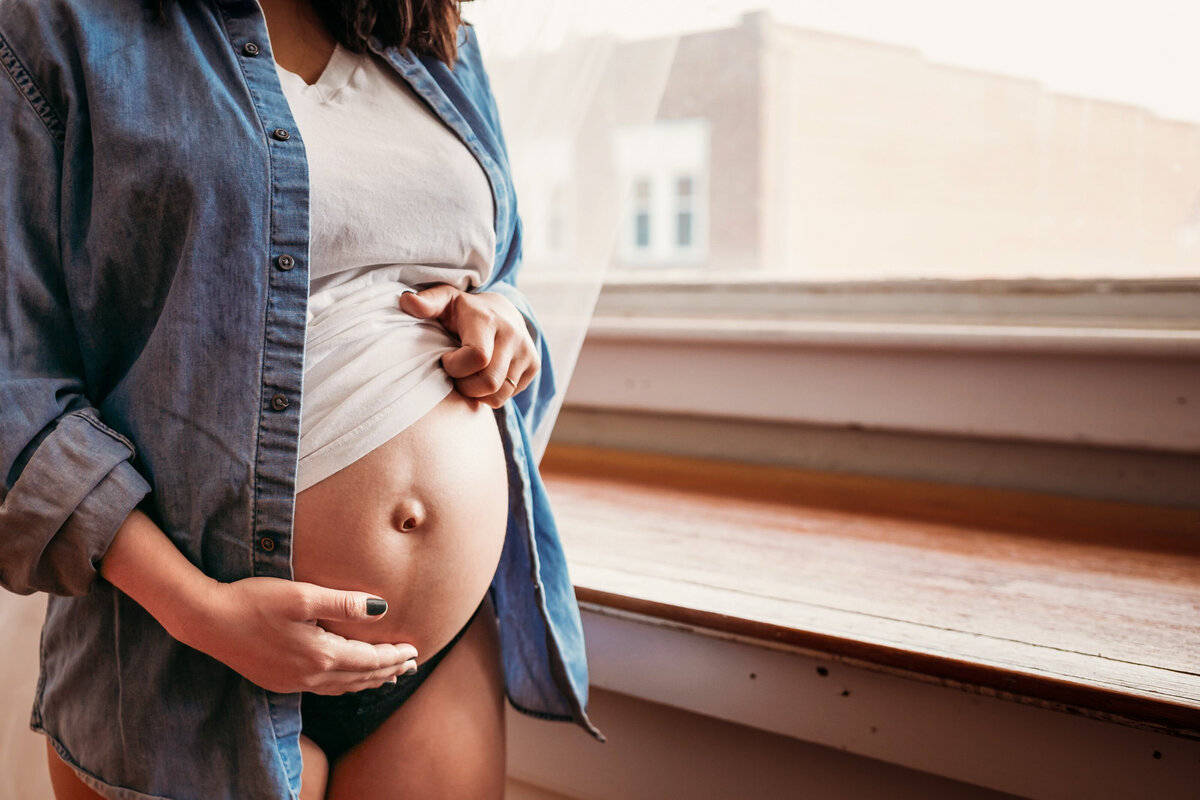 a close up of a woman in a white tee and denim shirt  exposing her belly in front of a window