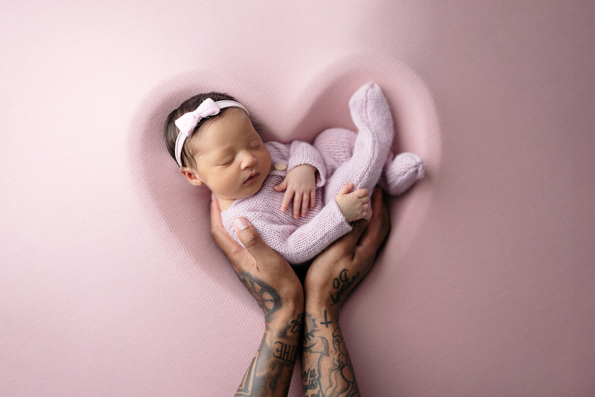 Newborn baby girl wearing a pink knit footie is held by her dad while laying inside of a heart shape during her newborn photo shoot in Memphis.