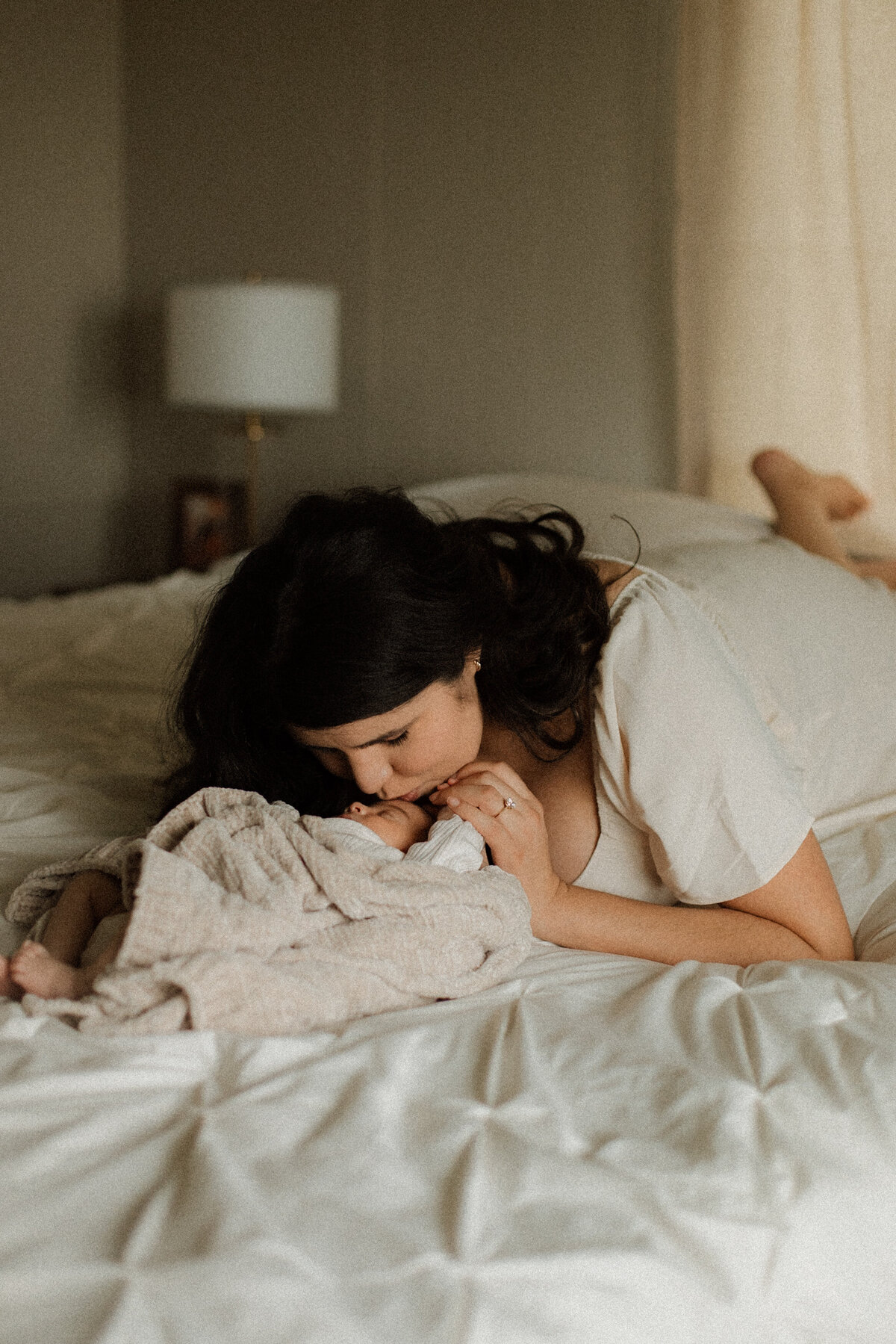 Mother laying on the bed, kissing her newborn wrapped in a soft blanket, in a peaceful bedroom setting.