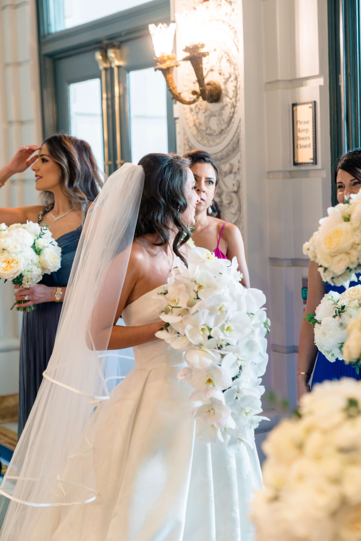 A bride in a white gown holds a cascading white bouquet, surrounded by three bridesmaids holding smaller bouquets. They stand indoors near ornate walls and large windows, engaged in conversation.