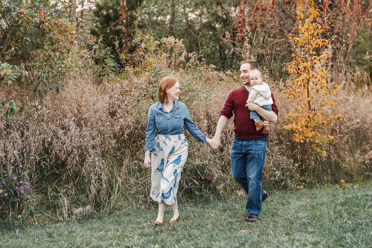 Strolling amidst tall grass and vibrant autumn foliage, a couple walks hand in hand while the man carries their baby. Captured by a talented Toronto family photographer