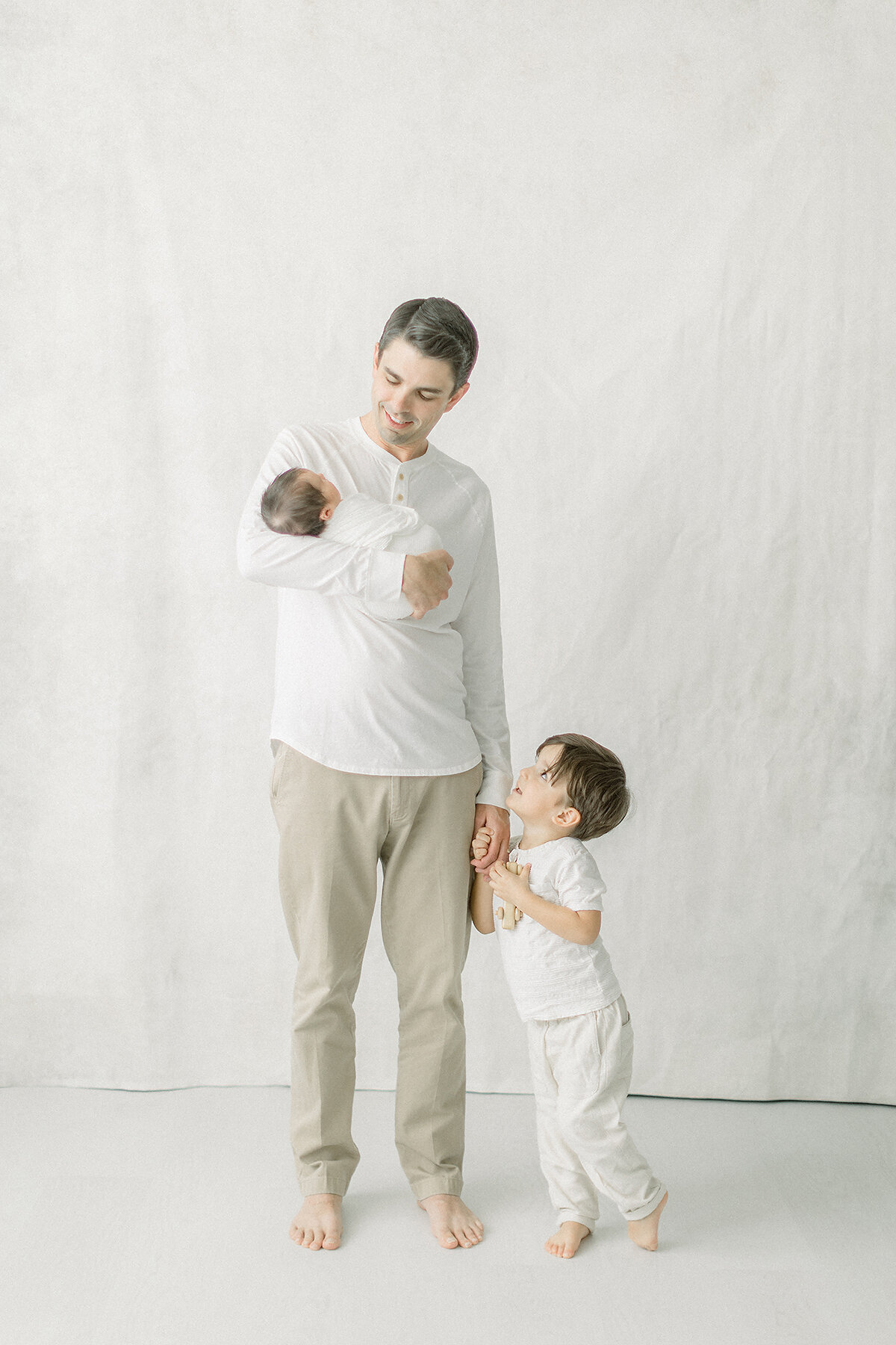 A light filled family portrait of a dad holding his newborn baby with his toddler son by his side as they take their new family photos in a photography studio by Dallas/Fort Worth, TX.