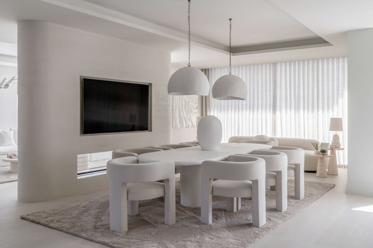 A modern dining room with a minimalist aesthetic, featuring a large oval table surrounded by six curved white chairs. Two large, white dome-shaped pendant lights hang above the table. The room is decorated in neutral tones with a plush, light-colored rug underneath the table and chairs. Behind the table, a large television recessed in the cream wall.