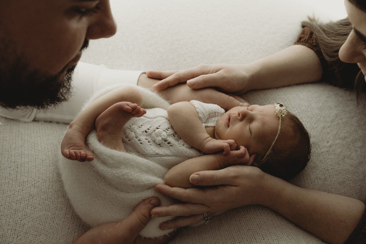Adults holding a newborn baby who is wrapped in a white blanket and wearing a knitted outfit and headband during their Wollongong motherhood session. The baby is sleeping peacefully.