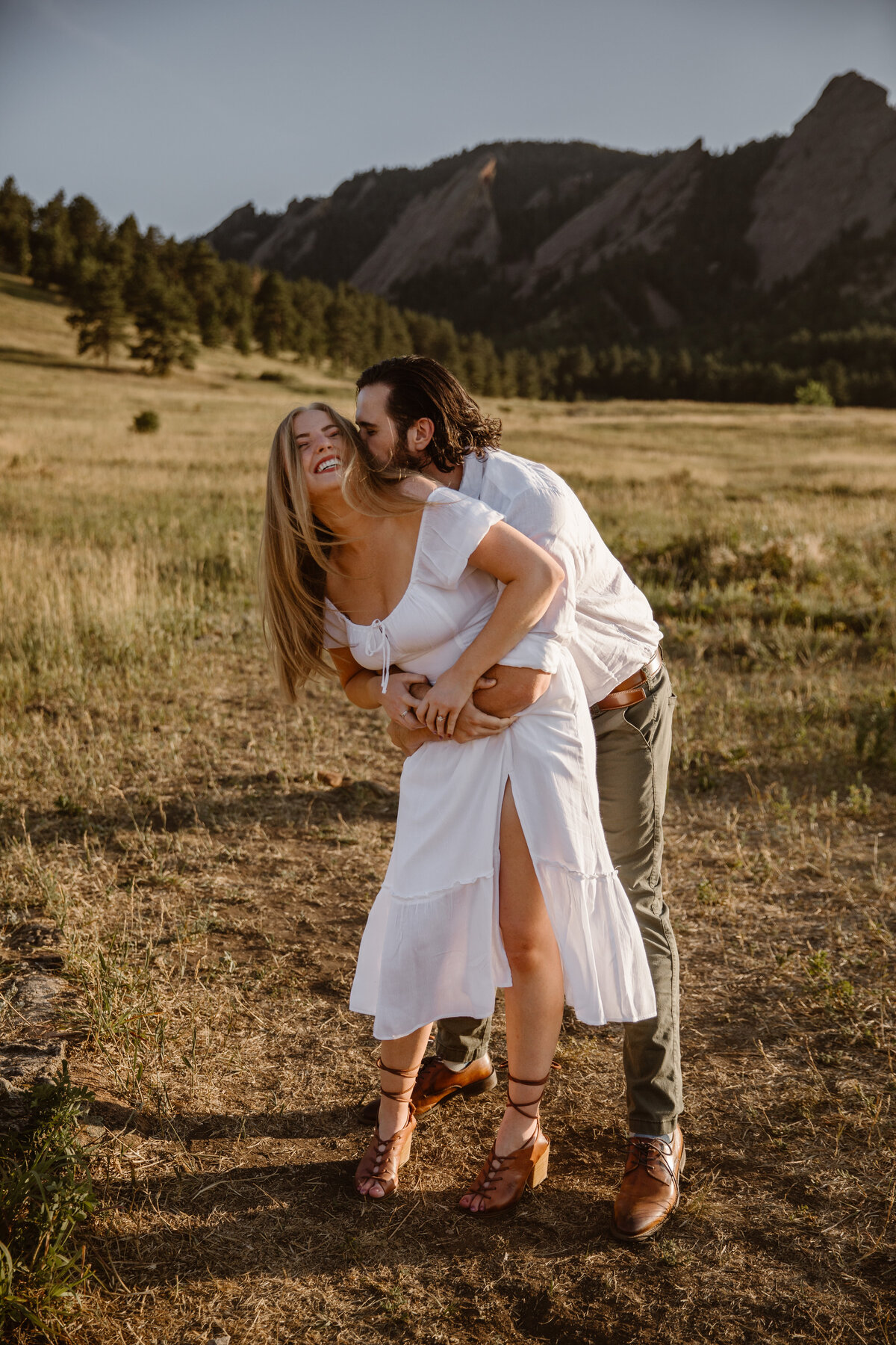 A man wraps his arms around his laughing partner from behind in a golden field with the Flatirons mountains in the background.